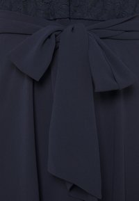 Navyblaues Kleid mit einem Spitzenoberteil, fließendem Stoff und einer großen Schleife an der Taille. Glatte Textur mit strukturierter Silhouette.