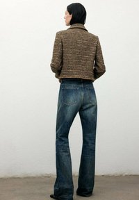 Chaqueta de tweed marrón con cuello, diseño corto; combinada con jeans acampanados de color azul oscuro. Tela texturizada y prominentes bolsillos traseros visibles.