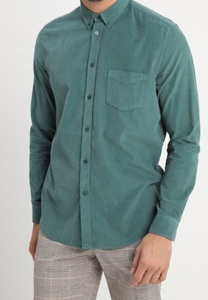 Homme portant une chemise verte en velours côtelé boutonnée avec une poche poitrine et un pantalon à carreaux, debout devant un fond uni.