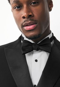 Black tuxedo with satin lapels, white shirt, and a textured black bow tie. Notable details include black buttons and a sleek, tailored fit.
