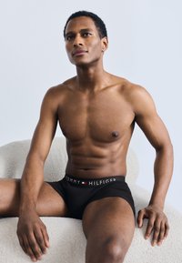 Μαύρα εσ ώρουχα boxer με λογότυπο στη ζώνη που γράφει "TOMMY HILFIGER." Το μοντέλο έχει γυμνασμένο σώμα και κάθεται σε μια υφασμάτινη καρέκλα ανοιχτού χρώματος.
