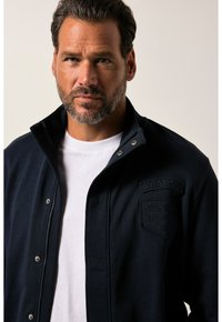 Giacca blu navy con colletto alto, cerniera frontale e bottoni a pressione. Dettaglio del logo dell'aviazione ricamato sul petto sinistro.