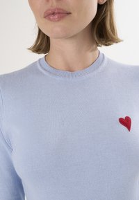 Maglione lavorato a maglia di un azzurro chiaro con scollatura rotonda, caratterizzato da un ricamo a forma di cuore rosso sul lato sinistro. Texture morbida, design aderente.