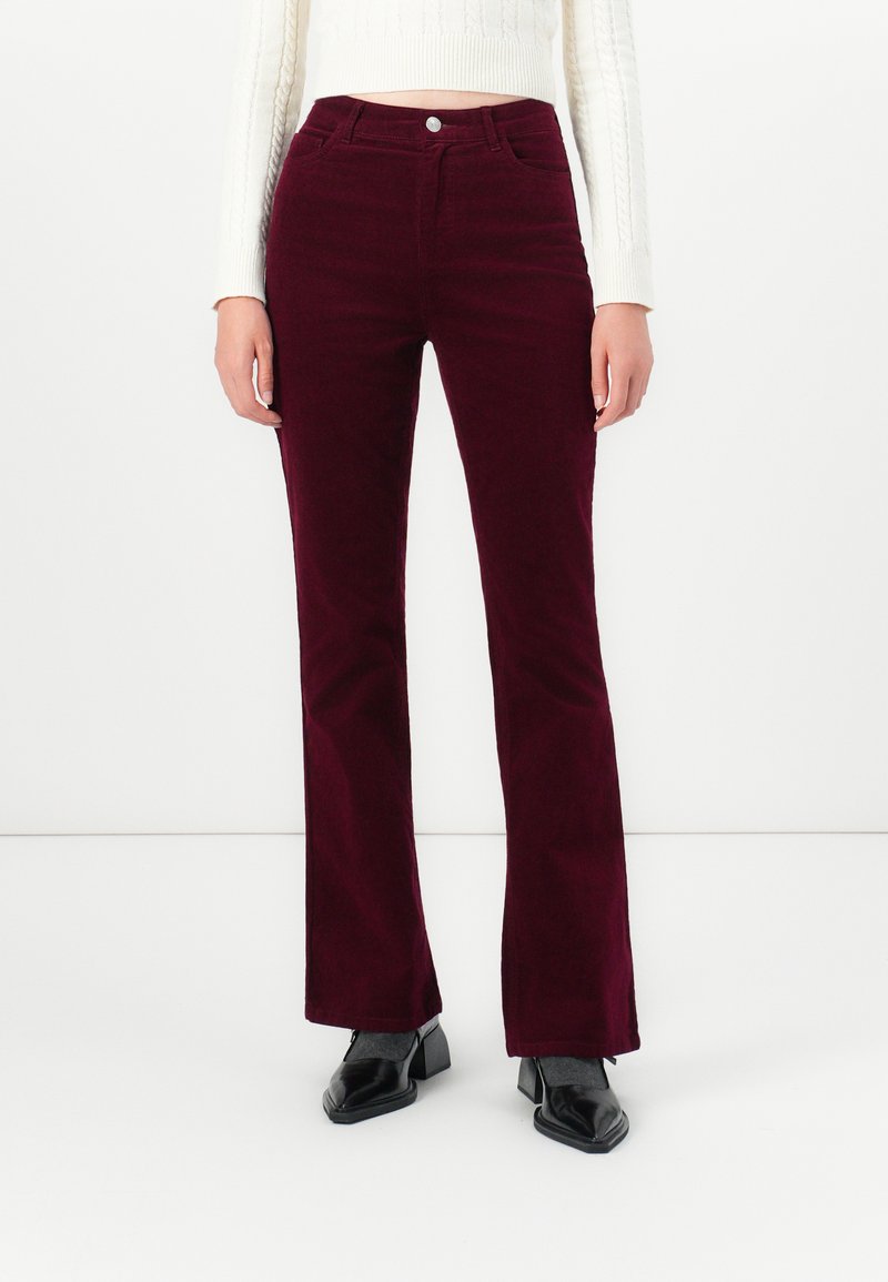 Vrouw in hooggewaisted, donkerburgundy corduroy broeken gecombineerd met zwarte hakken en een witte cropped trui.