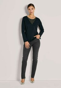 Schwarzes Langarmoberteil mit V-Ausschnitt und verziertem Kragen; kombiniert mit dunkelgrauen, schmal geschnittenen Jeans und hellgrauen High Heels.
