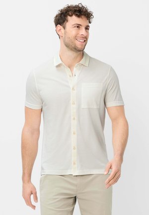 Lächelnder Mann mit lockigem Haar, der ein beige kurzärmeliges Hemd mit Brusttasche und hellbeige Hose trägt.