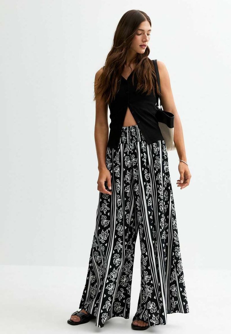 Haut sans manches noir avec une fermeture à boutons, associé à un pantalon large à rayures noires et blanches avec des motifs floraux. Sandales simples noires.