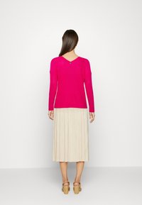 Svetr s výstřihem do V a dlouhými rukávy z jasně růžové pleteniny; na zadní straně je detail s knoflíkem. Sluší mu světle plisovaná midi sukně.