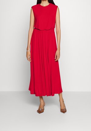 Abito lungo rosso senza maniche con vita arricciata, tessuto liscio e gonna fluente. Modella che indossa scarpe marroni con punta a punta e dettagli con fiocco.