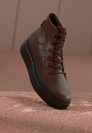 Baskets montantes en cuir marron avec semelle noire épaisse et lacets, positionnées debout sur une surface beige texturée et un arrière-plan.