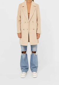 Abrigo de lana beige de doble botonadura con solapas de muesca, dos botones y bolsillos laterales; combinado con unos jeans acampanados de denim desgastado en azul claro y zapatillas blancas.