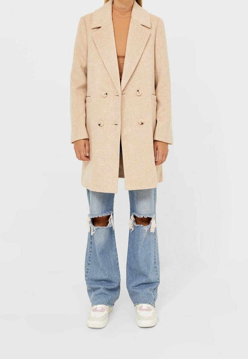 Abrigo de lana beige de doble botonadura con solapas de muesca, dos botones y bolsillos laterales; combinado con unos jeans acampanados de denim desgastado en azul claro y zapatillas blancas.