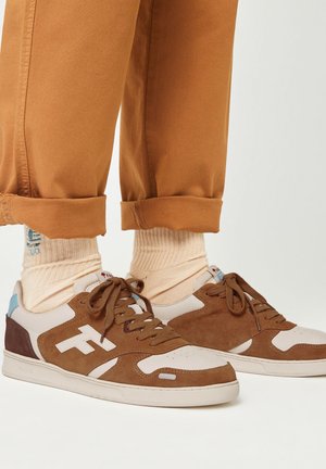 Baskets marron et beige avec un dessus en daim et cuir, présentant un logo blanc, un bout rond et une semelle en caoutchouc plate. Portées avec des chaussettes beiges.