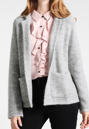 Kvinde iført en lysegrå åben-front strikket cardigan over en lyserød bøjlekantet skjorte med knapper og sorte bukser.