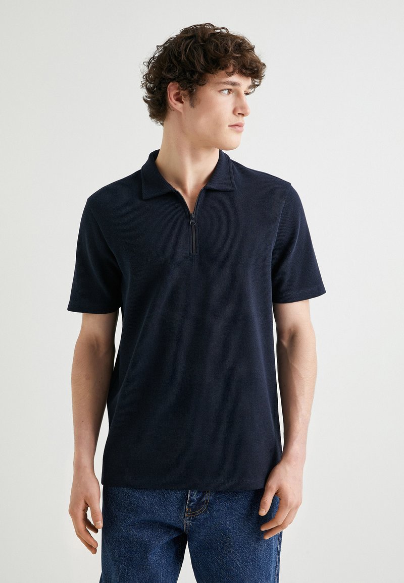 Marinblå kortärmad polo-shirt med dragkedja vid kragen. Tillverkad av texturerat tyg, med avslappnad passform och en stilren design. Bärs tillsammans med jeans.