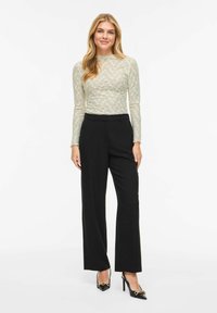 Top in pizzo verde chiaro a maniche lunghe abbinato a pantaloni neri a vita alta e décolleté nere con punte e dettagli dorati.