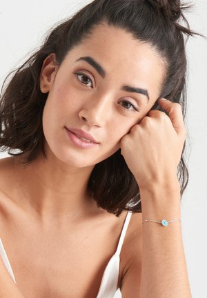 Silberarmband mit einer kleinen türkisfarbenen Scheibe und roten Akzenten, getragen an einem Handgelenk mit natürlichem Hautton. Einfaches, dünnes Kettendesign.