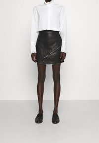 Camicia bianca a maniche lunghe con dettagli in pizzo e una gonna avvolgente nera in pelle. Scarpe nere senza lacci completano l'outfit su uno sfondo neutro.