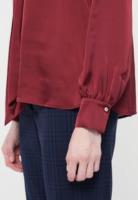 Blusa bordeaux con maniche lunghe, tessuto morbido, polsini arricciati e dettagli con bottoni, abbinata a pantaloni a quadri blu navy.