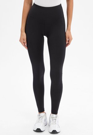 Person wearing black high-waist leggings and white athletic shoes, standing against a plain white background.