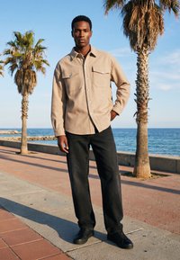 Homme portant une chemise beige boutonnée et un pantalon noir, debout sur une promenade en bord de mer avec des palmiers et l'océan en arrière-plan.