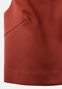 Un tissu couleur rouille présente une surface lisse et texturée avec une couture diagonale et un petit détail de poche angulaire. Bords nets et silhouette structurée.