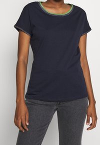 Námořnická modrá tričko s krátkým rukávem z bavlny s krkem lemovaným zeleným a stříbrným pruhem. Má uvolněný střih a jednoduché švy.