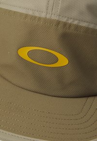 Khaki caps med teksturert overflate, med en fremtrende gul oval logo, strukturert kuppel og sydelige detaljer langs sk brim.