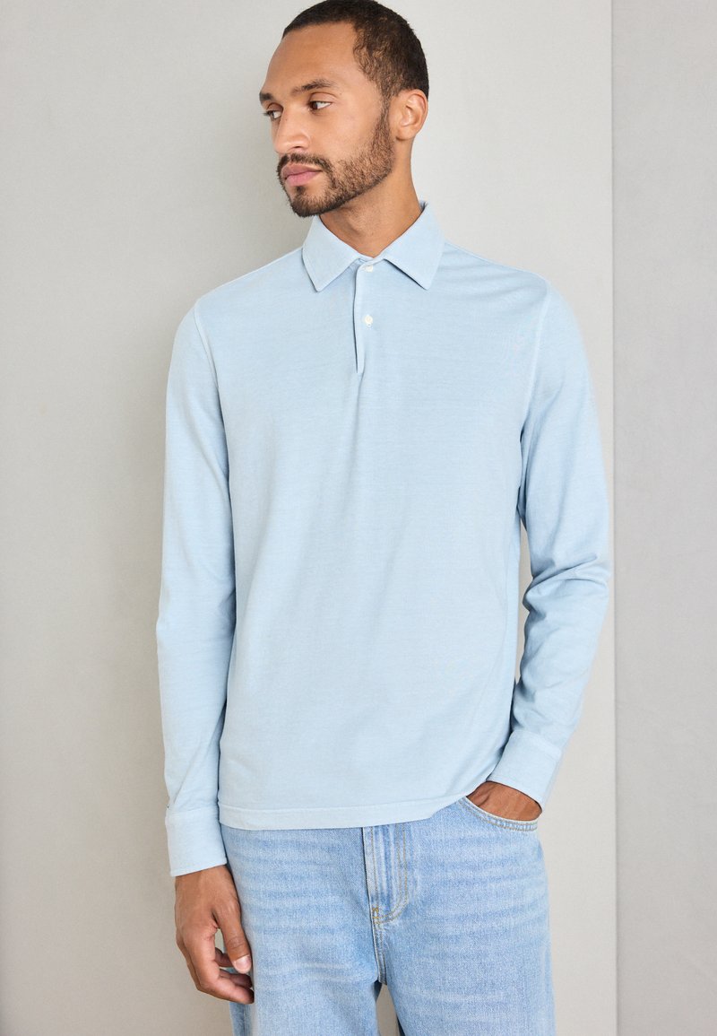 Mann med kort skjegg iført en lyseblå langermet polo-skjorte og lyse vaskede jeans, som ser til siden med den ene hånden i lommen.