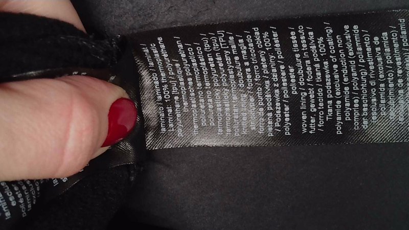 Etykieta na czarnej odzieży wykonanej z poliestru, szczegółowo opisująca materiały w wielu językach; teksturowana powierzchnia z błyszczącym wykończeniem, częściowo zasłonięta ręką.