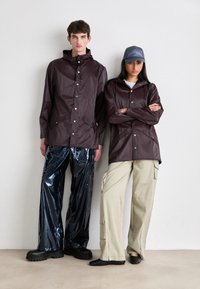 Burgunderfarget vanntett jakke med hette og to frontlommer, sammen med glinsende svarte bukser. Beige cargo-bukser med glidelås og detaljer. Sorte støvler.