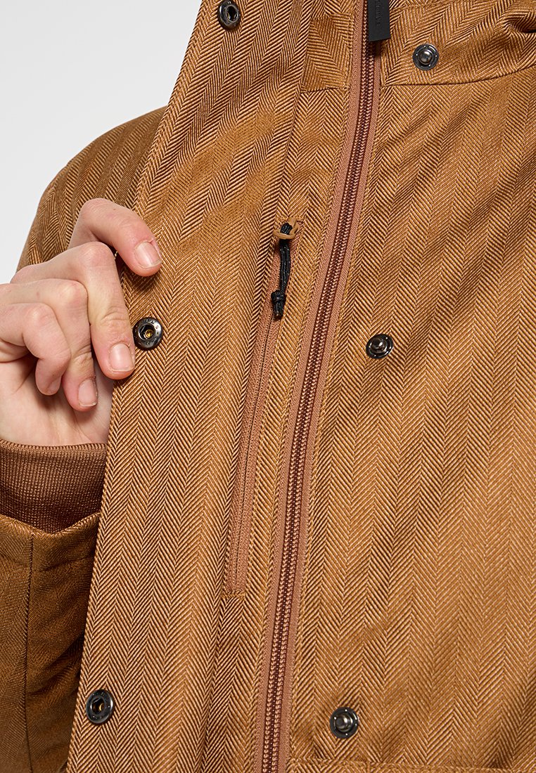 Braune Fischgrätenjacke mit Reißverschluss und Druckknopfverschlüssen, die eine strukturierte Oberfläche aufweist und von einer Hand am Kragen gehalten wird.