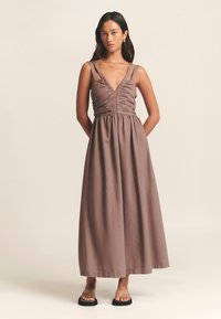 Maxikjole i brunt stof med gathered bodice, dyb V-hals og tynde justerbare stropper. Flade sandaler fuldender outfittet.