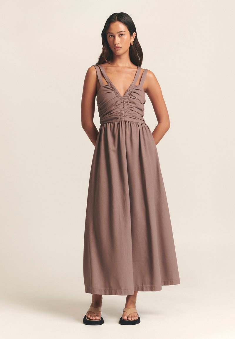 Maxikjole i brunt stof med gathered bodice, dyb V-hals og tynde justerbare stropper. Flade sandaler fuldender outfittet.