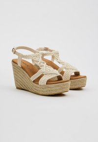 Sandalias de cuña con correas de crema trenzadas, plantilla marrón texturizada y tacón envuelto en yute. Correa ajustable en el tobillo con cierre de hebilla.
