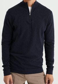 Marinblå zip-up tröja i mjukt tyg, med hög krage och långa ärmar med ribbade manschetter, kombinerad med ljusa byxor.