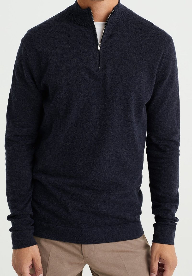 Marinblå zip-up tröja i mjukt tyg, med hög krage och långa ärmar med ribbade manschetter, kombinerad med ljusa byxor.
