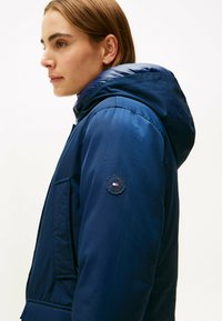 Giacca puffer blu navy con cappuccio, dotata di una patch con logo sulla manica sinistra e un tessuto liscio e leggero. Tasca frontale visibile.