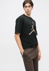 Czarna bawełniana koszulka z kamuflażowym logo Air Jordan, zestawiona z brązowymi dresami, przedstawiona w perspektywie z góry.