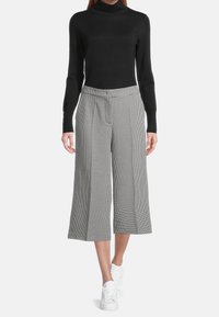 Žena v černém dlouhém roláku, černobílých kostkovaných širokých kalhotách v cropped střihu a bílých teniskách na bílém pozadí.