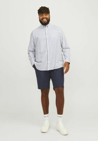 Striped button-up shirt in white and navy, paired with navy shorts. Model wears white sneakers and white socks. Simple and casual look.