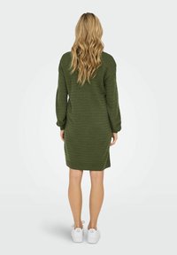 Robe tricotée vert olive avec manches longues, poignets et ourlet côtelés. La texture est douce, avec de subtiles rayures verticales. Associée à des baskets blanches.