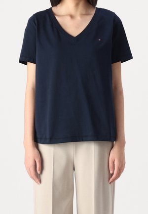 Personne portant une chemise à manches courtes bleu marine avec col en V et un pantalon beige clair, debout devant un fond uni.