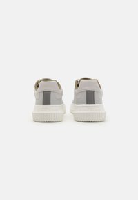 Sneakers grigie chiare con accenti beige, caratterizzate da una suola testurizzata, collo imbottito e logo sul tallone posteriore. Parte superiore in pelle liscia.