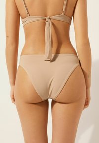 Beige bikini-set med knytning i ryggen och högskurna underdelar. Släta, elastiska tyger med minimalistisk design och inga synliga mönster eller detaljer.
