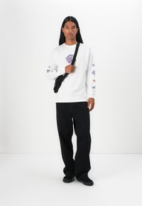 White long-sleeve shirt with colorful graphic designs on sleeves, paired with loose black pants and black shoes, featuring a crossbody bag.
