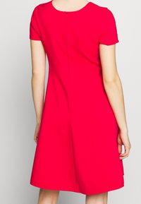 Takana näkymä henkilöstä, joka on pukeutunut polviikään, lyhythihaiseen, kirkkaan punaiseen mekkoon, jossa on sauma vyötäröllä ja takana vetoketjukiinnitys.