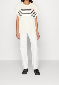 Lyhythihainen kerma t-paita mustilla vaakaraitkuilla ja punaisella "Hilfiger"-logolla; yhdistetty valkoisiin suoralahkeisiin housuihin ja mustiin tasapohjakenkiin.