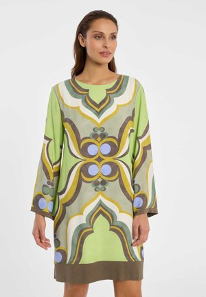 Frau trägt ein locker sitzendes, langärmliges Kleid mit kräftigen, symmetrischen abstrakten Mustern in Grün, Gelb, Braun, Weiß und Blau.