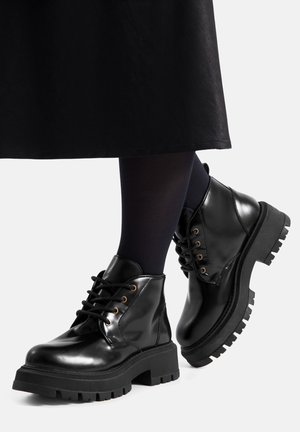 Botines de cuero patentado negro con suelas gruesas y resistentes, punta redonda y cordones. Presentan ojales de latón y suelas exteriores de goma texturizada.